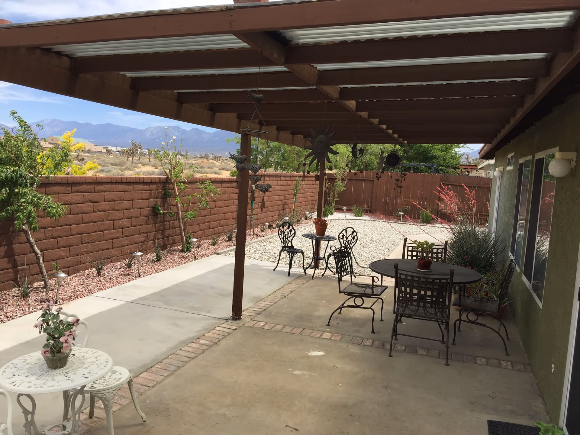 Outdoor patio area with a wooden pergola overhead, several metal tables and chairs, potted plants, and a brick wall enclosing the space. Desert landscape and mountains are visible in the background.