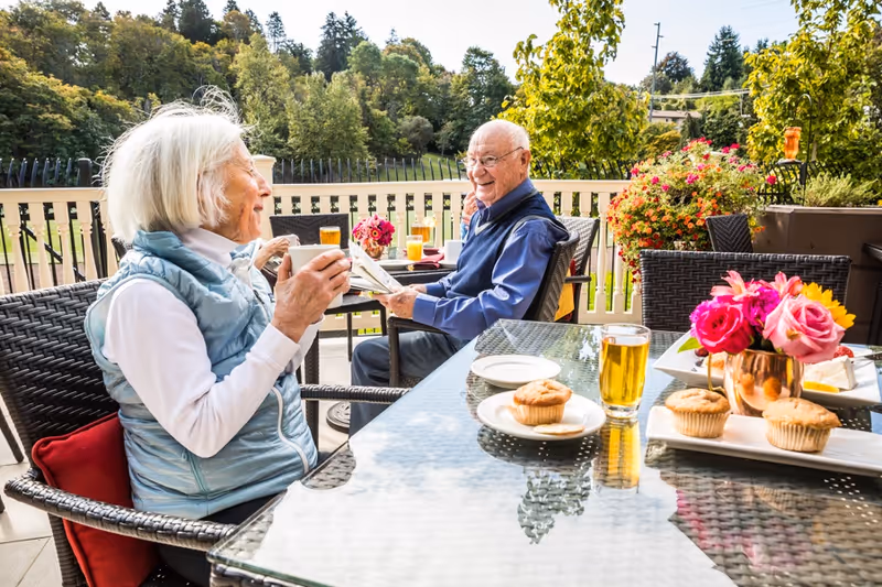 Two elderly people sitting at an outdoor patio table enjoying drinks and muffins. The woman on the left is holding a cup and smiling, while the man on the right is holding a newspaper and smiling back. The table has plates with muffins, glasses of juice, and a vase with colorful flowers. The background shows greenery and trees under a clear sky.