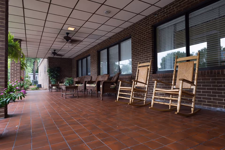 Covered brick porch with rocking chairs, wicker seating, potted plants, and ceiling fans along a building exterior.