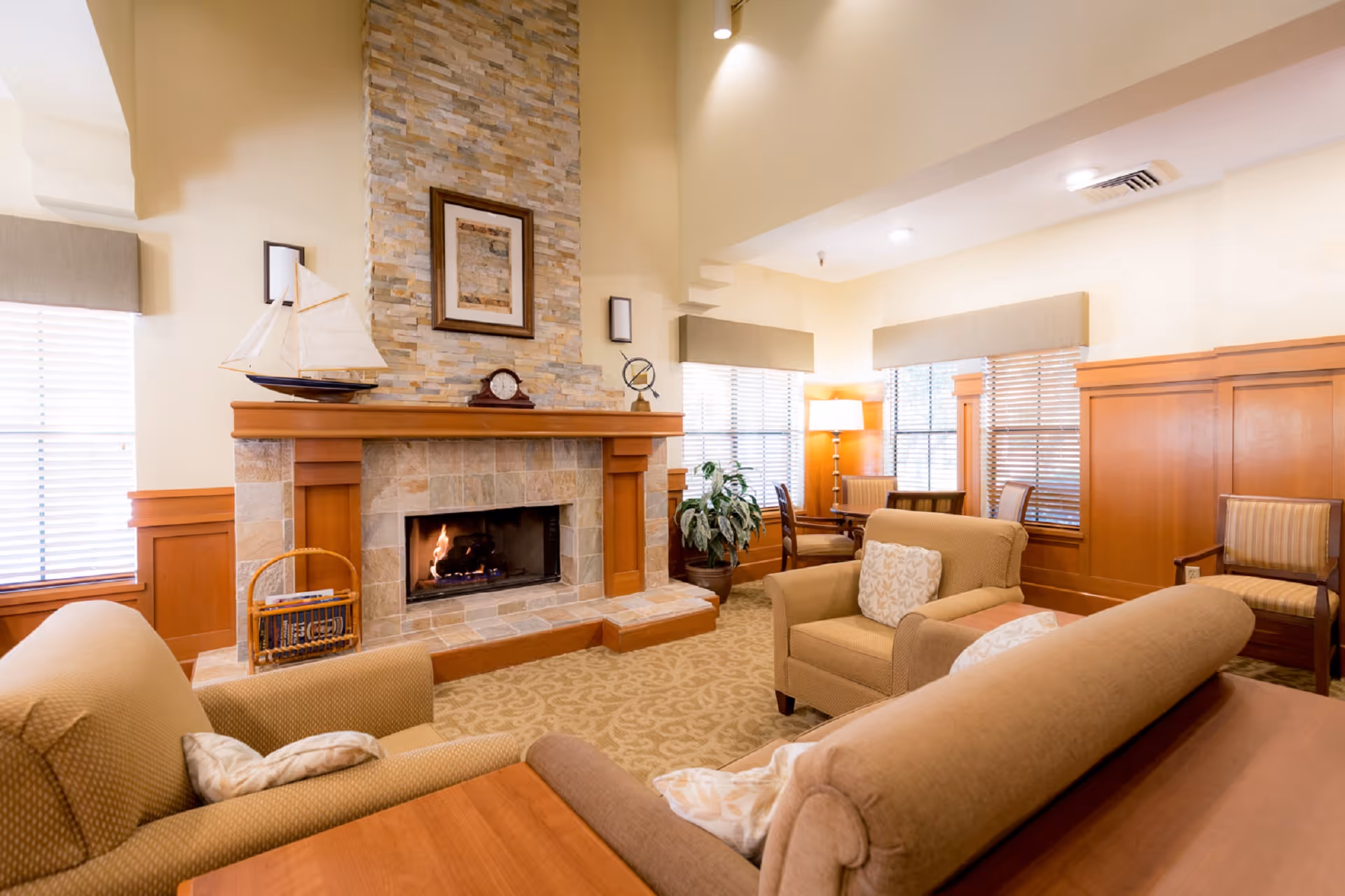 A cozy living room area with beige upholstered armchairs and a sofa arranged around a stone fireplace with a wooden mantel. The mantel is decorated with a model sailboat, a clock, and a small globe. The room has large windows with blinds, wooden paneling on the walls, a patterned carpet, and a floor lamp in the corner providing warm lighting.