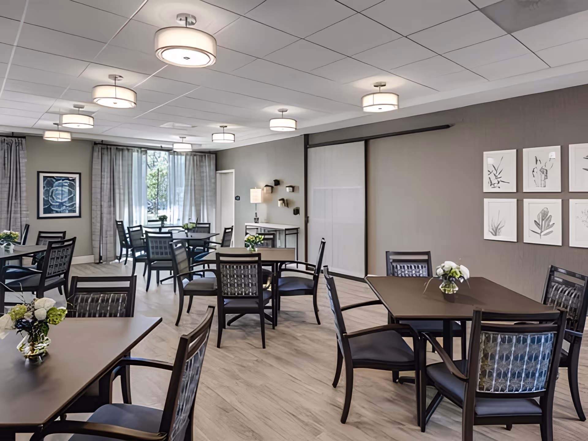 A well-lit dining room with multiple dark wood tables and chairs arranged neatly. Each table has a small floral centerpiece. The room features light-colored walls, large windows with sheer curtains allowing natural light, modern ceiling lights, and framed artwork on the walls.