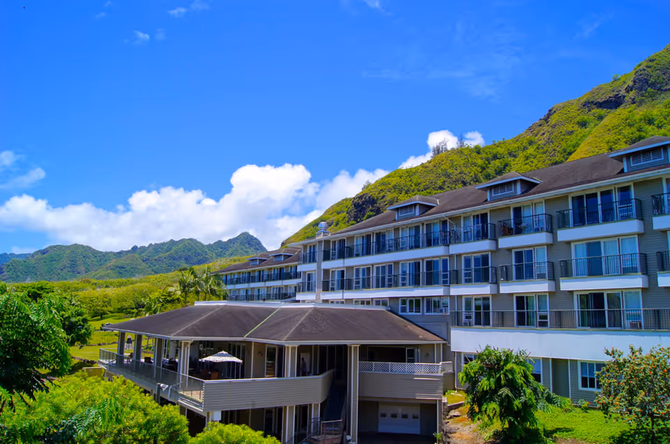Exterior view of a multi-story senior living facility named Brighton Rehabilitation, surrounded by lush greenery and mountains under a bright blue sky with some clouds.