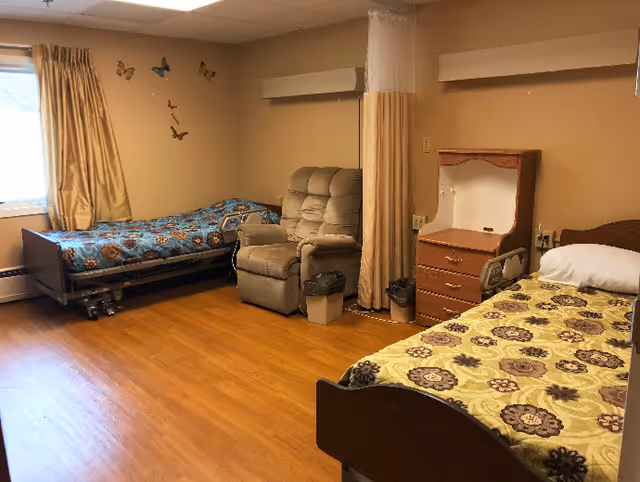 Shared senior living bedroom with two beds, a recliner, a nightstand, and a privacy curtain.