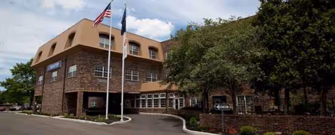 A brick senior living facility exterior with flags, trees, and an accessible entrance.