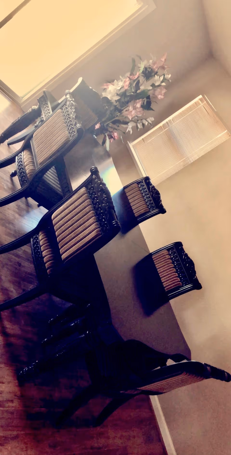 A dining room with a dark wooden dining table and six matching chairs with upholstered seats and backs. A floral arrangement in a vase is placed on the table. The room has wooden flooring and a window with closed blinds.