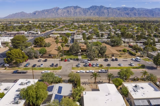 Aerial view of a residential neighborhood with single-story buildings, parking lots, and streets, set against a backdrop of a mountain range under a clear blue sky.