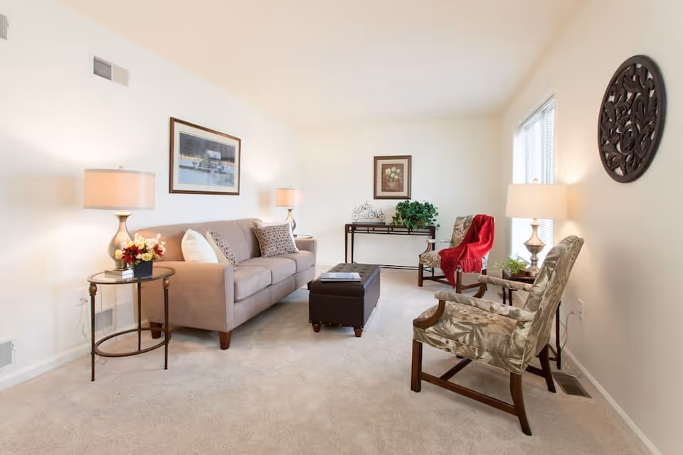 A bright and cozy living room with a beige sofa adorned with pillows, two patterned armchairs, a dark ottoman in the center, and two side tables with lamps and decorative plants. The walls are decorated with framed artwork and a circular wooden wall piece. Large windows allow natural light to fill the room.