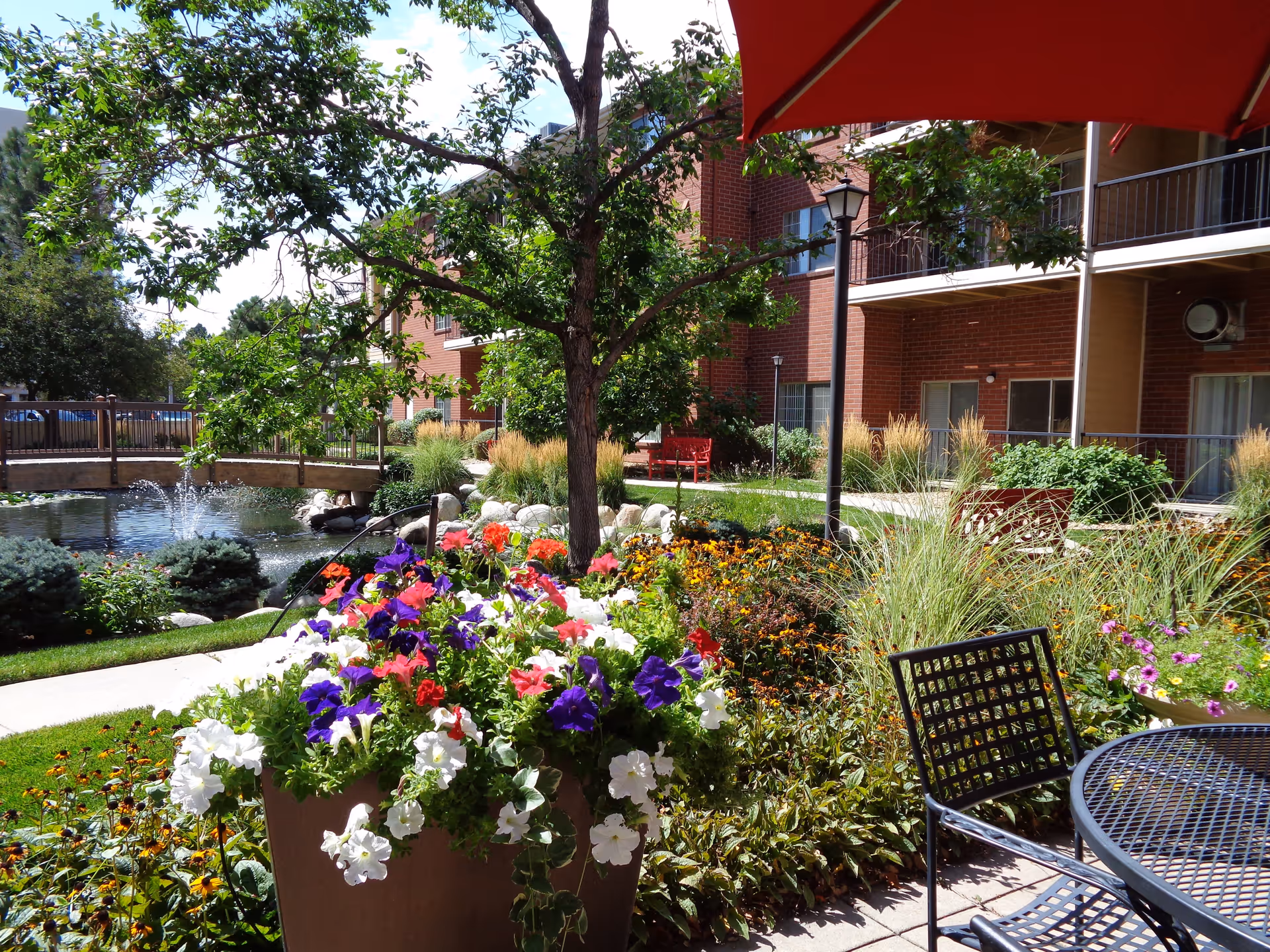A vibrant outdoor garden area at Springbrooke Retirement featuring colorful flowers in a large planter, a tree, a small pond with a fountain, a wooden bridge, and a brick building with balconies in the background. There is also a black metal table and chair set under a red umbrella in the foreground.