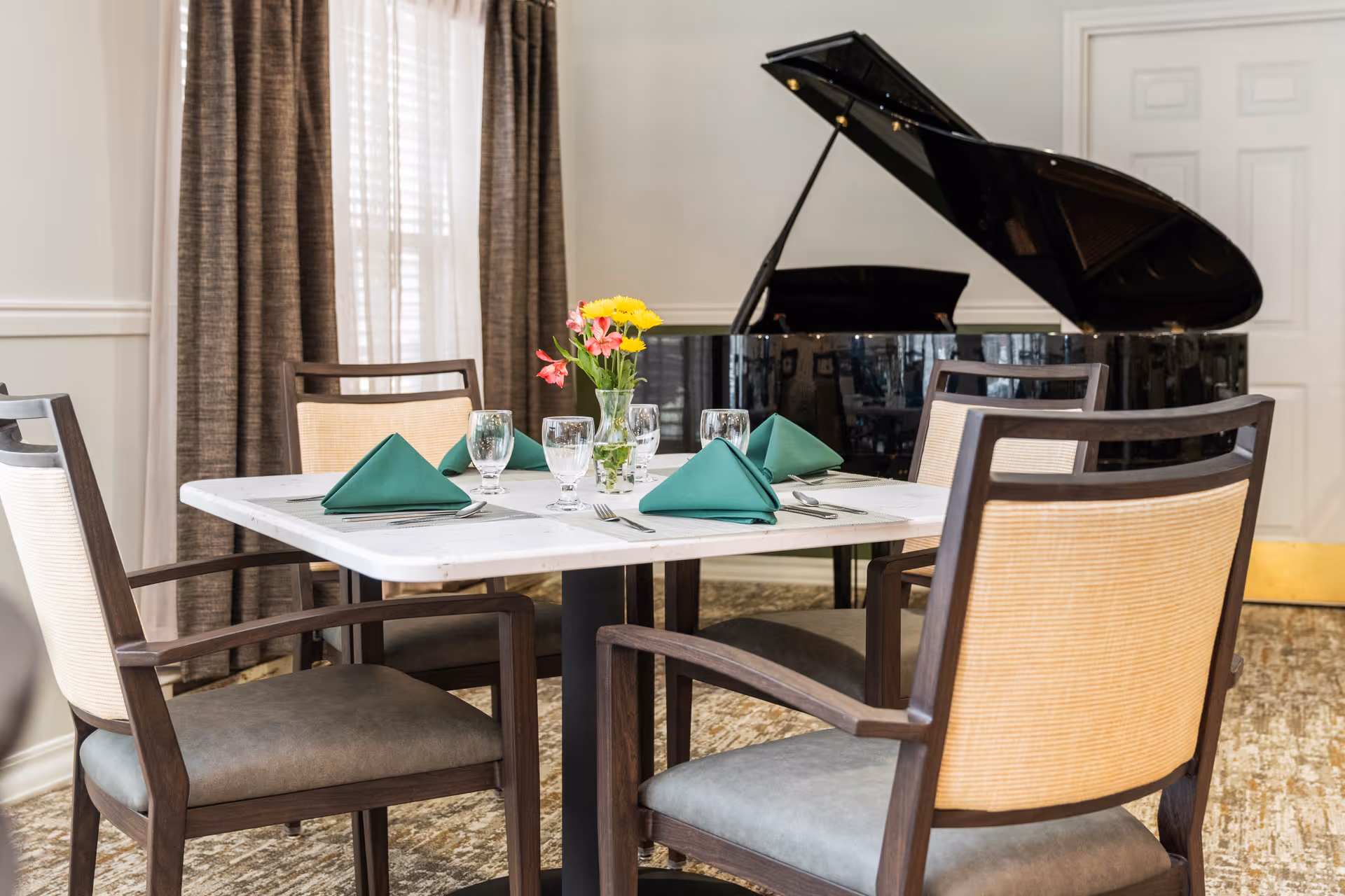 Dining table set with four chairs, green napkins, glassware and a small vase of flowers, with a grand piano in the background.
