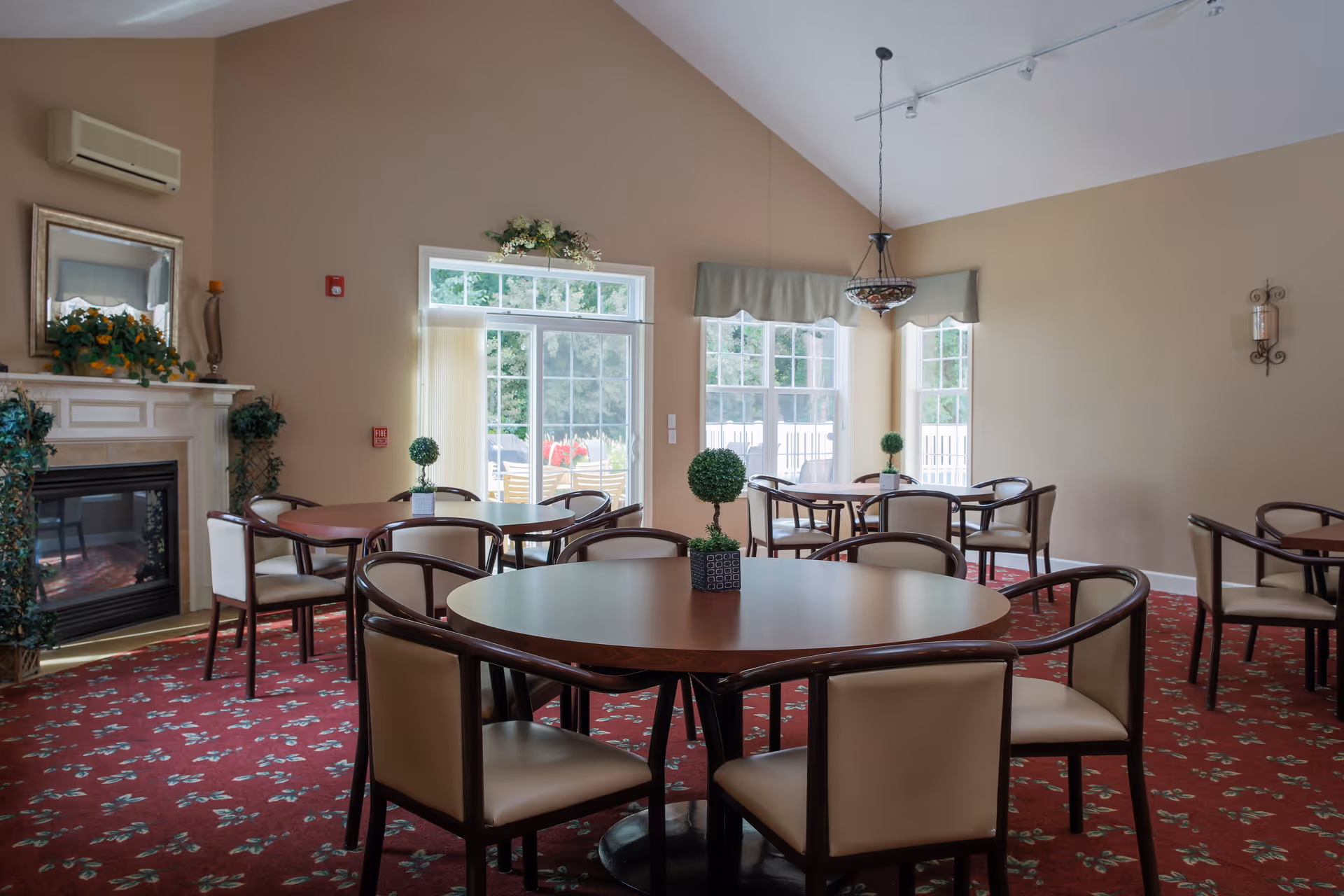 A dining room with round wooden tables and cushioned chairs arranged neatly on a red carpet with a floral pattern. The room has large windows letting in natural light, a fireplace with decorative plants and a mirror above it, and a hanging light fixture from the ceiling.