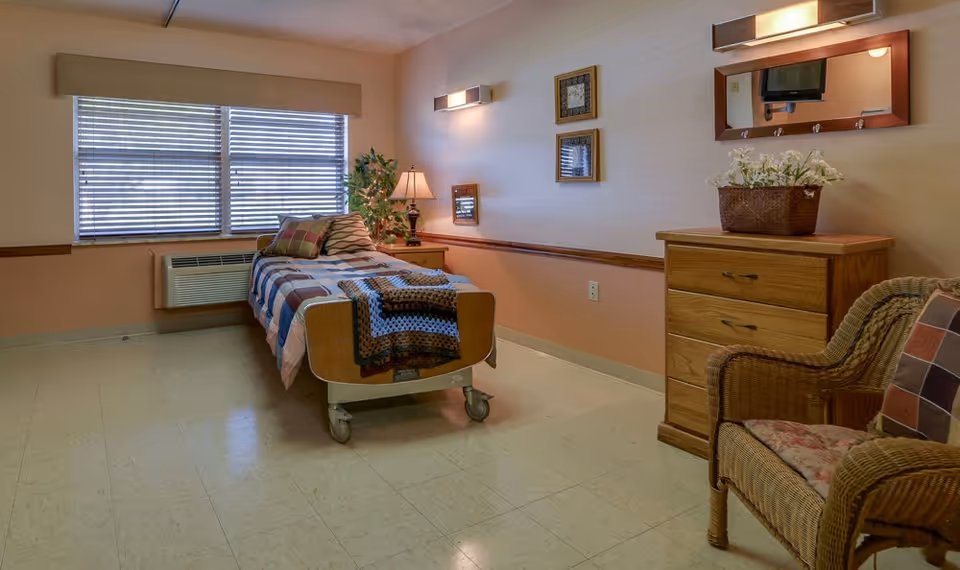 A neatly arranged nursing center bedroom with a single bed covered in a patchwork quilt, a wooden dresser with a flower basket on top, a wicker chair with a cushion, a lamp on a small table, and a large window with blinds letting in natural light.