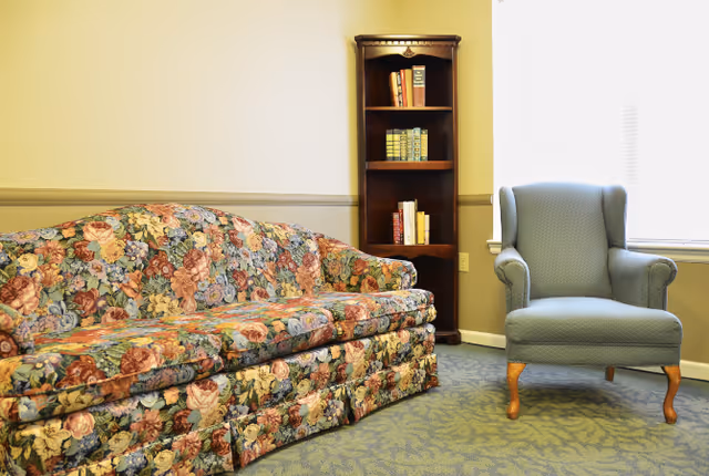 A cozy sitting area with a floral patterned sofa, a blue upholstered armchair, a wooden bookshelf with books, and a window with blinds letting in natural light.