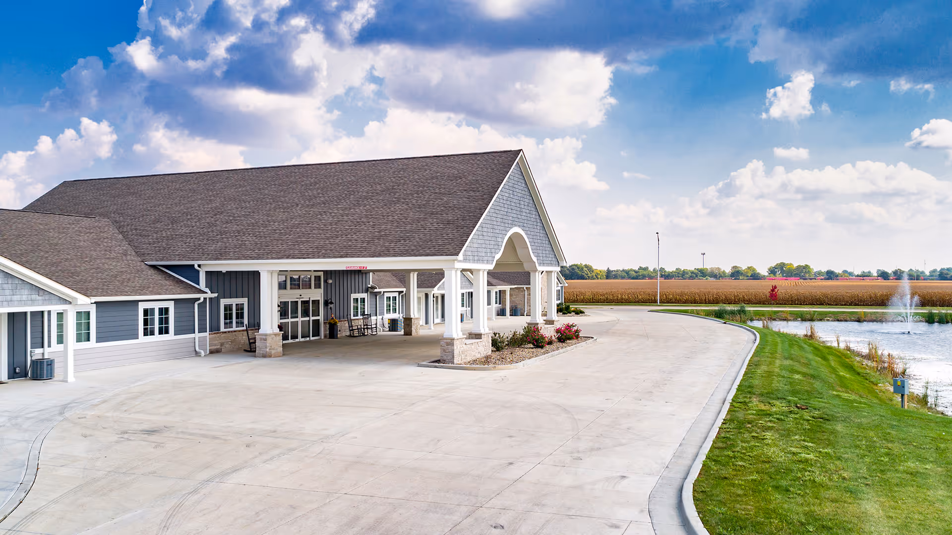 Exterior view of Villas of Holly Brook Assisted Living & Memory Care building in Streator, IL, showing a large covered entrance with columns, a driveway, green lawn, a pond with a fountain, and a partly cloudy sky.