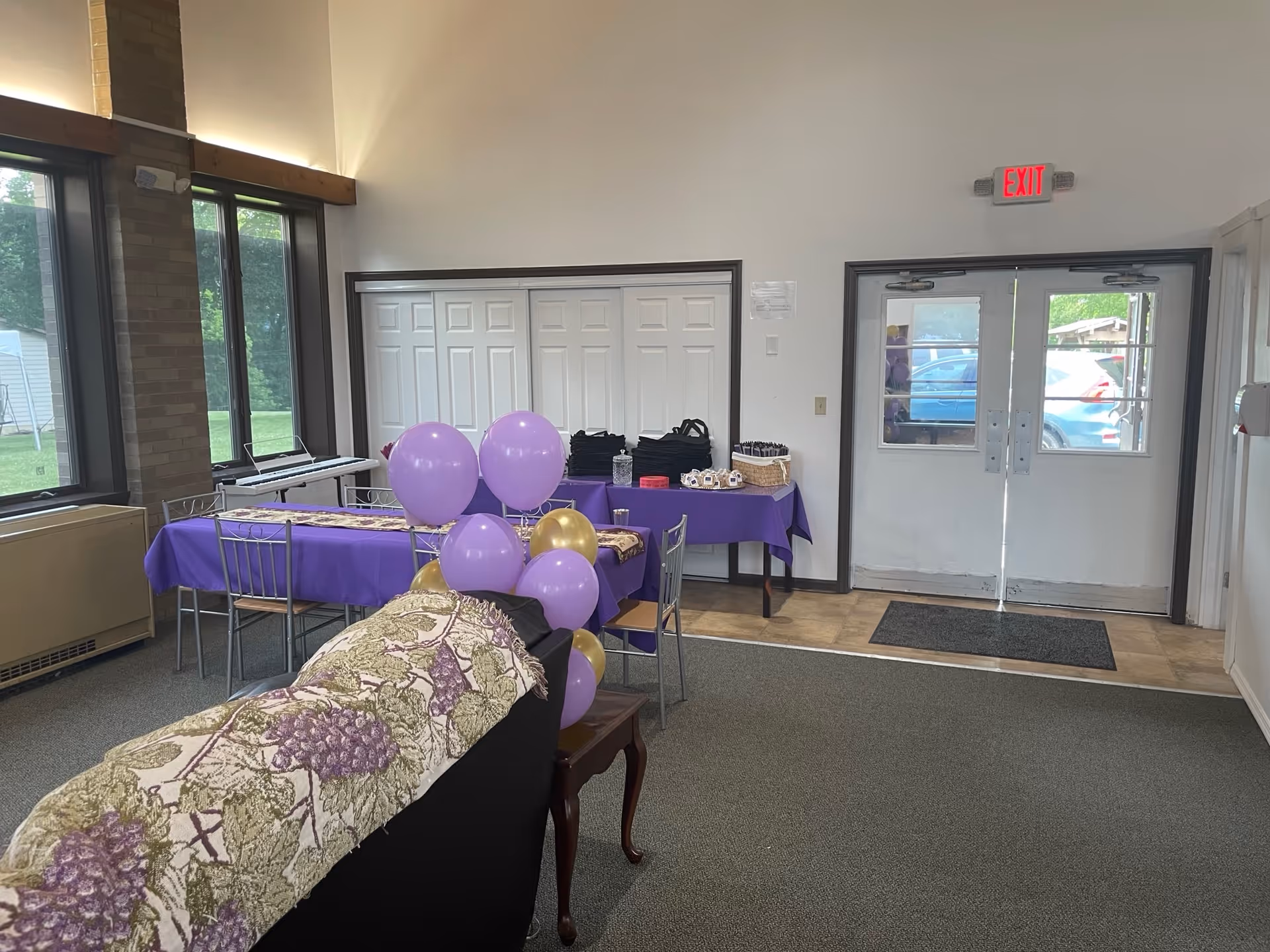 Community room with tables covered in purple tablecloths, purple and gold balloons, chairs, and double exit doors.