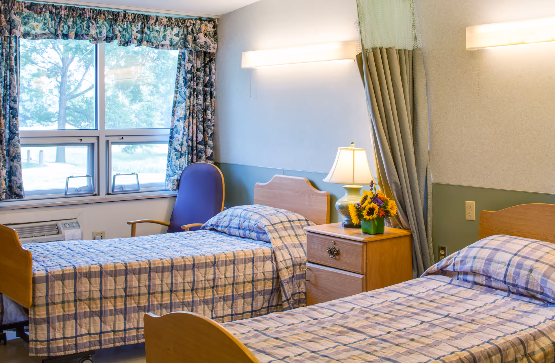 A bright room with two single beds covered in plaid bedspreads. Between the beds is a wooden nightstand with a lamp and a small vase of sunflowers. A blue chair is positioned near a window with floral curtains, and soft wall lights illuminate the room.