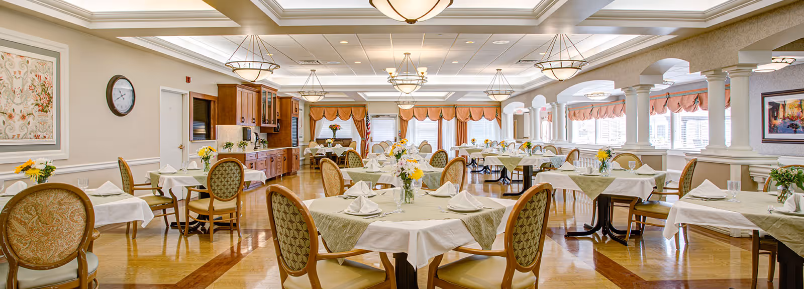 Spacious, well-lit dining room with round tables set with white tablecloths, floral centerpieces, cushioned chairs, and large windows.