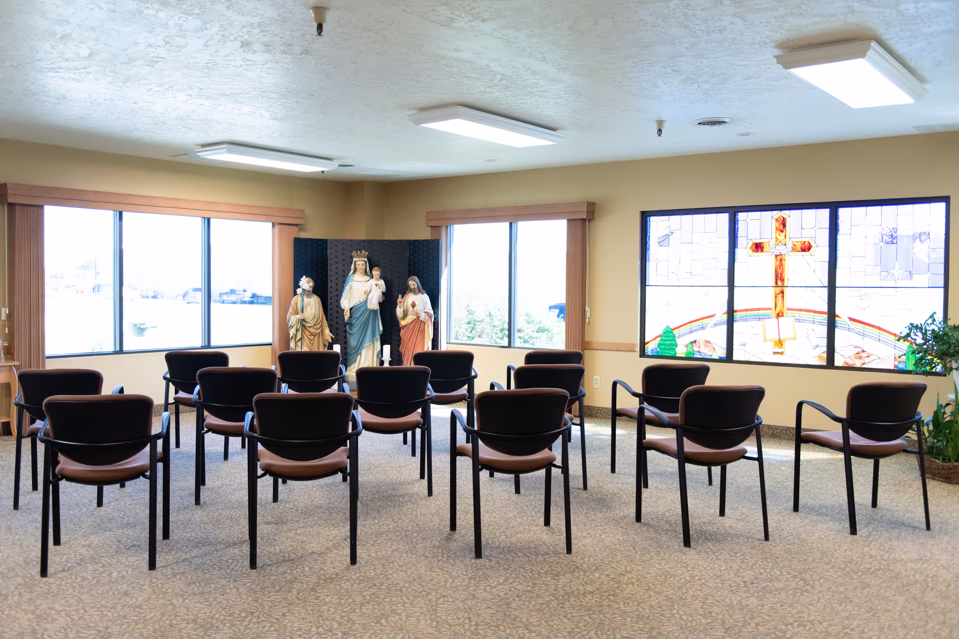 A small chapel or prayer room with rows of empty chairs facing religious statues and a stained glass window featuring a cross and a rainbow. The room has large windows letting in natural light and beige walls.