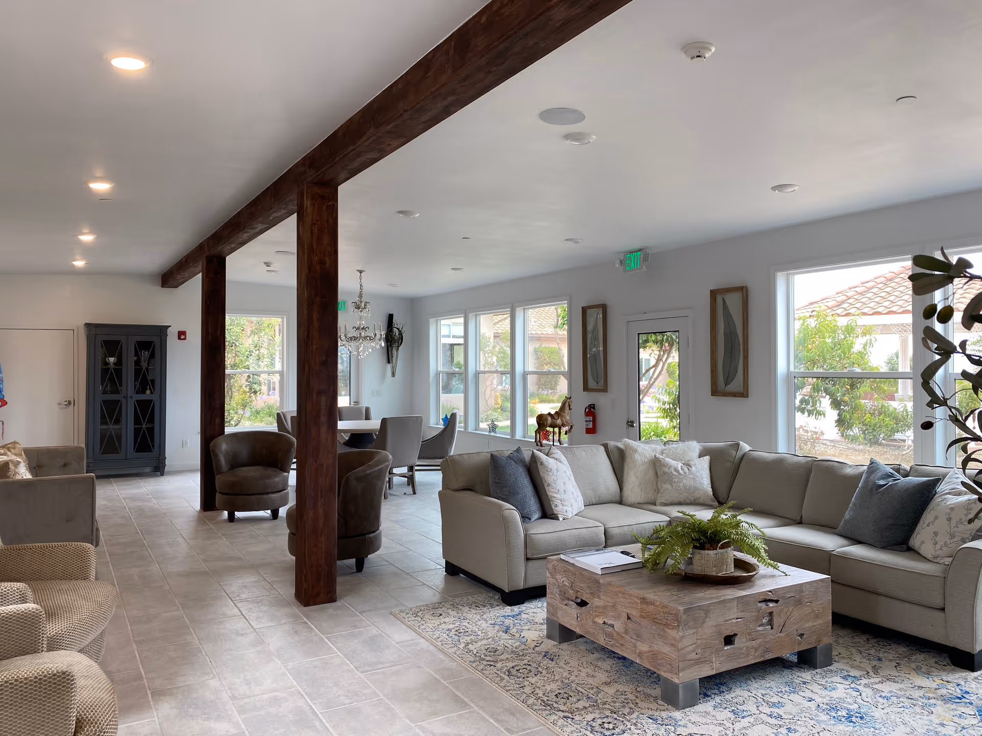 A spacious and well-lit living room area in a senior living facility with a large beige sectional sofa adorned with various cushions, a rustic wooden coffee table with a plant and books on it, several armchairs, and large windows letting in natural light. The room features tiled flooring, wooden support beams, framed artwork on the walls, and a chandelier hanging over a dining table in the background.