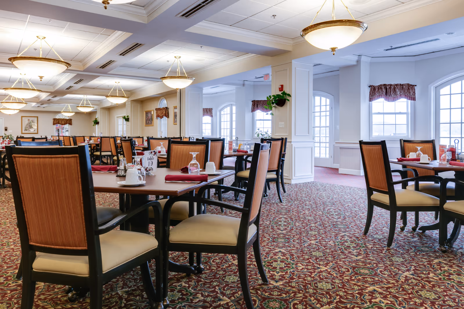 A spacious dining room in a senior living facility with multiple tables and chairs arranged neatly. Each table is set with cups, glasses, napkins, and condiments. The room features patterned carpet, large windows with valances, and ceiling lights providing warm illumination.