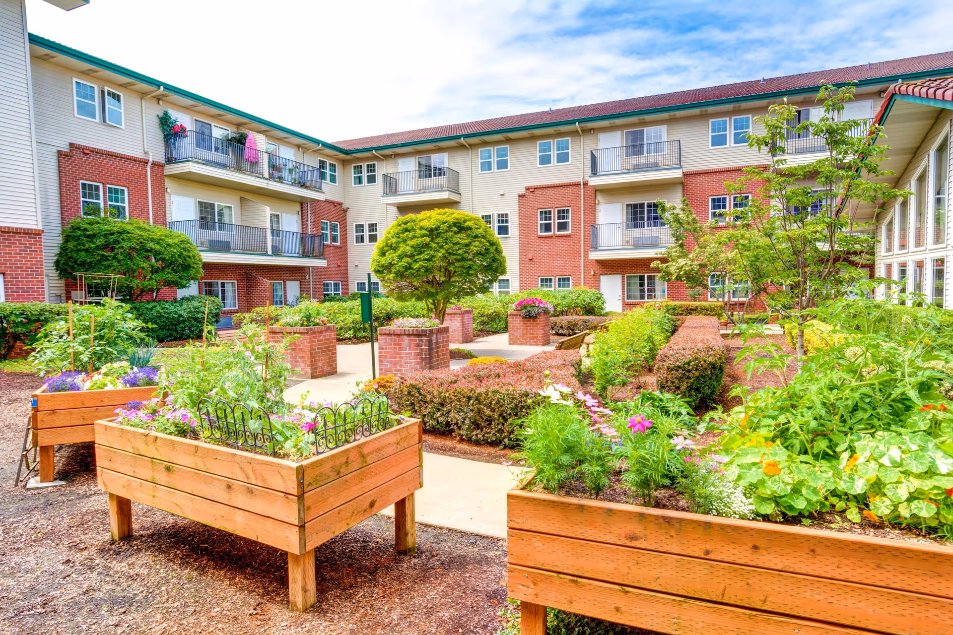 Outdoor courtyard garden area at Rosewood Park facility with raised wooden planter boxes filled with various flowers and plants, neatly trimmed bushes, and small trees. The courtyard is surrounded by a multi-story residential building with balconies and windows, under a partly cloudy sky.