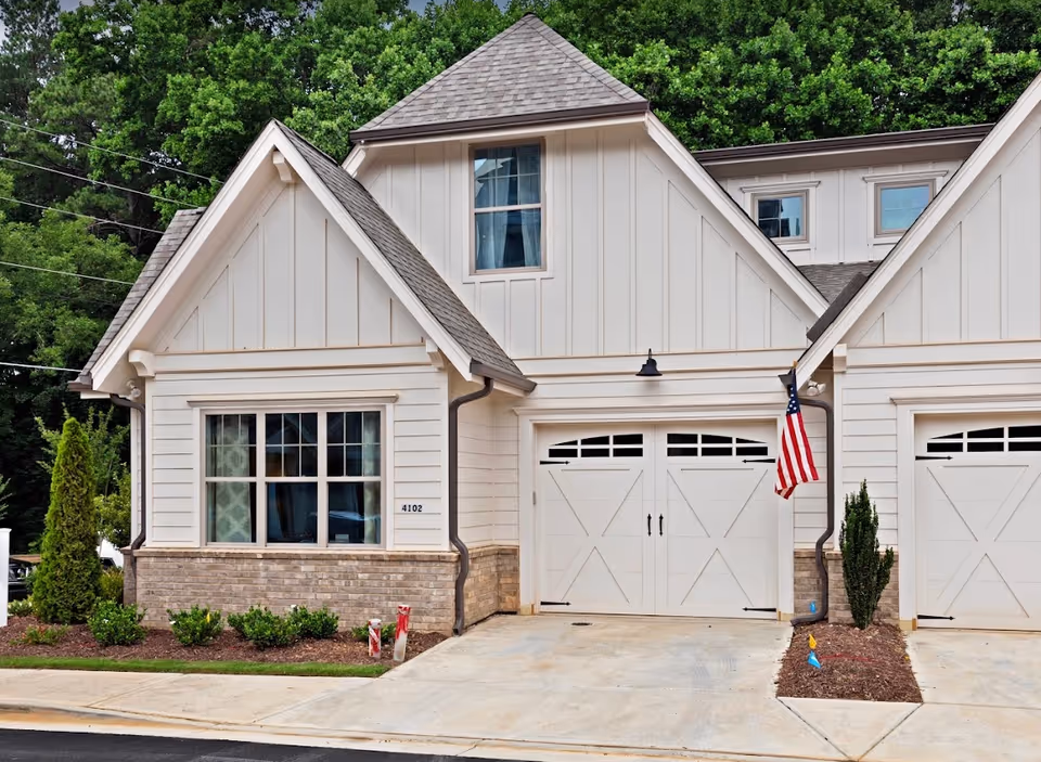 Exterior view of a modern residential building with white siding and brick accents, featuring a garage door with decorative windows and an American flag mounted on the wall. There are small shrubs and a tree planted in the landscaped area next to the driveway.