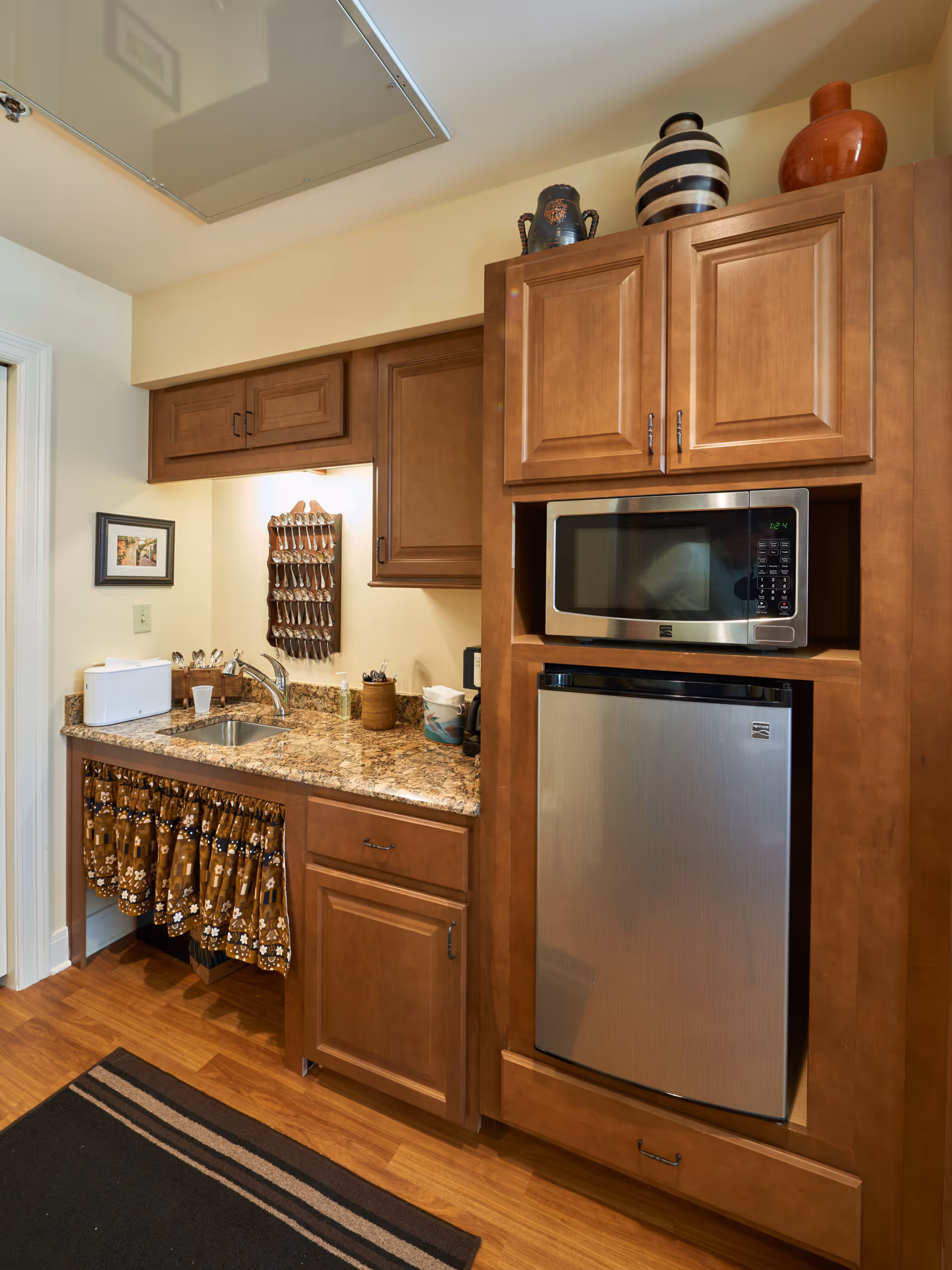 Small kitchenette with wooden cabinets, a granite countertop and sink, a microwave over a compact stainless refrigerator, and decorative jars on top.