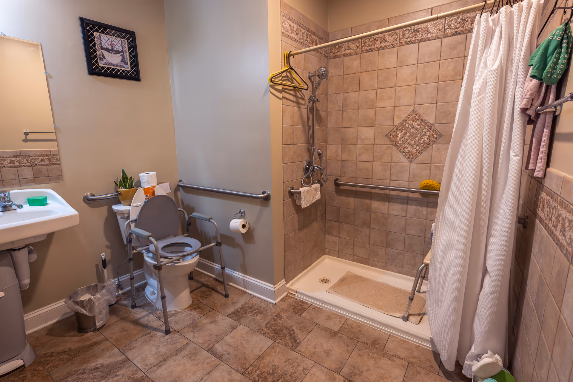 A senior-friendly bathroom with beige tiled floor and walls. It features a toilet with safety rails and a raised seat, a white pedestal sink, a walk-in shower with grab bars, a shower chair, and a white shower curtain. There is a small plant and toilet paper on the toilet tank, a framed picture on the wall, and a trash bin near the sink.