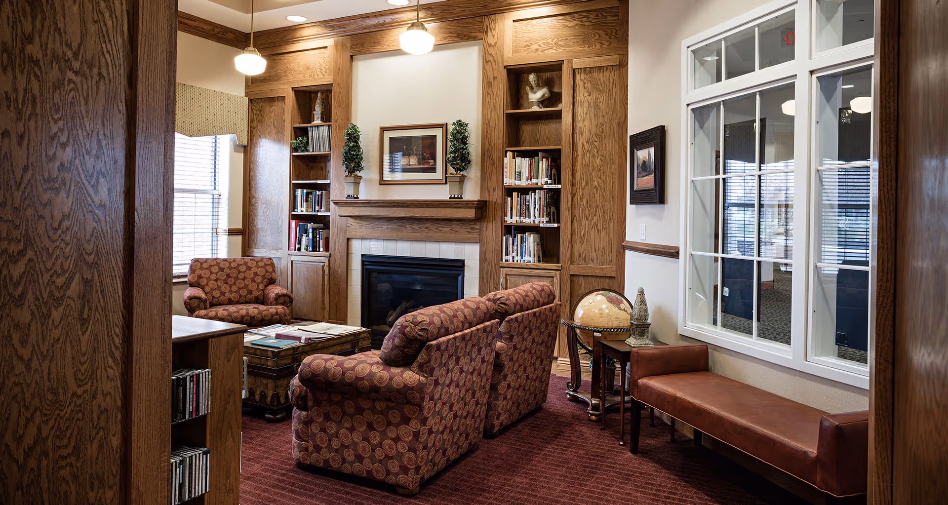 A cozy living room area with patterned armchairs and a sofa arranged around a wooden coffee table. The room features built-in wooden bookshelves flanking a fireplace with a framed picture above it. There are two small potted plants on the mantel. A leather bench sits against the wall under a large window with white trim. The room has warm wood paneling and a red carpeted floor.