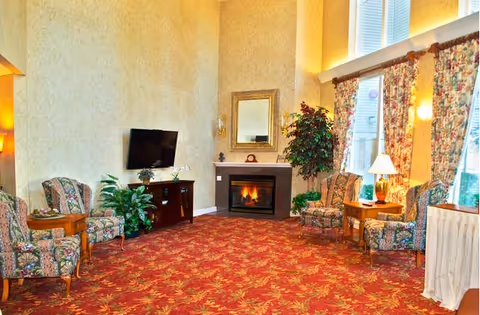 Bright common sitting room with floral upholstered chairs, a red patterned carpet, a TV, a fireplace, and tall windows with floral curtains.