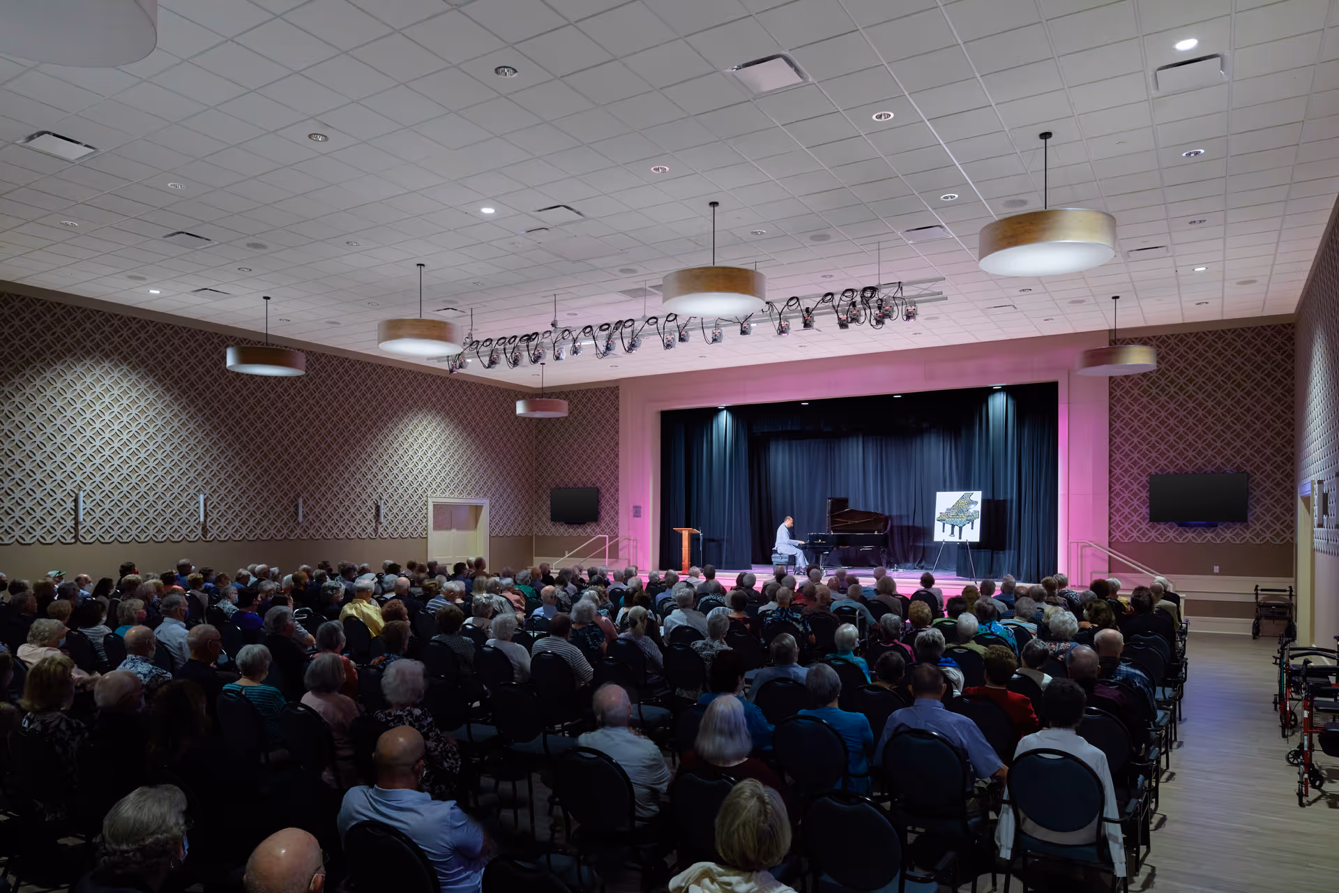 A large audience seated in rows of chairs watching a pianist performing on a stage in a spacious auditorium with patterned walls and ceiling lights.