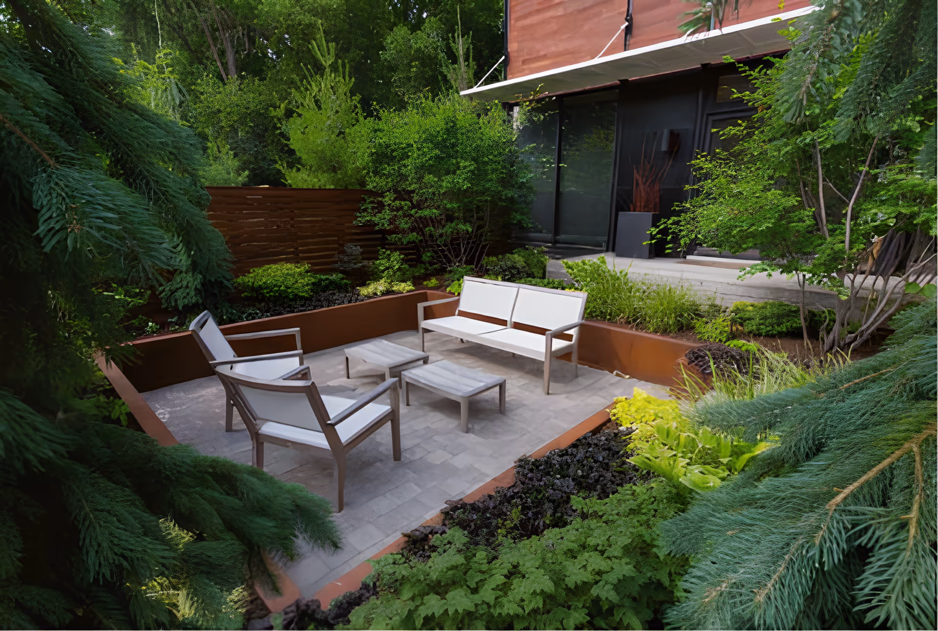 Outdoor patio area with modern white cushioned seating including a bench and two chairs around two small tables, surrounded by lush green plants and trees, adjacent to a building with large glass windows.