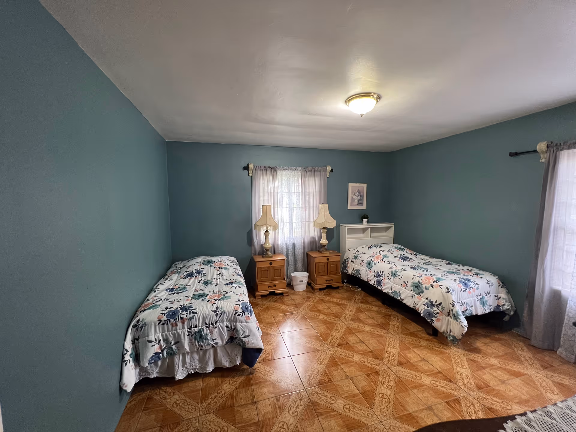 A bedroom with two single beds covered in floral bedspreads, each bed has a wooden nightstand with a lamp. The walls are painted teal, and there are two windows with sheer curtains allowing natural light to enter. The floor has a patterned tile design.