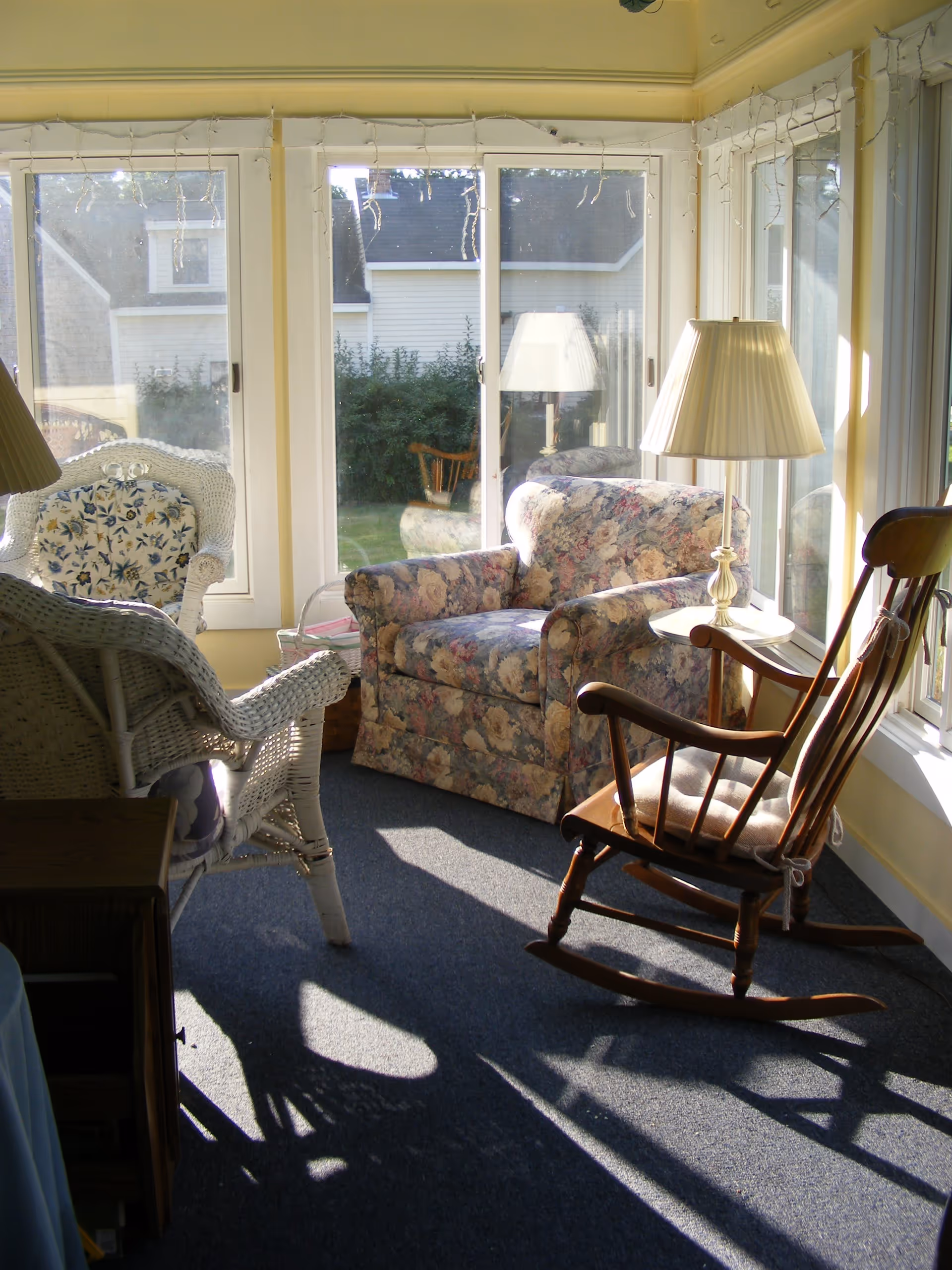 Sunlit sitting area with a floral upholstered armchair, a wooden rocking chair with a cushion, a white wicker chair with floral cushions, a small round table with a lamp, and large windows showing an outdoor view of houses and greenery.