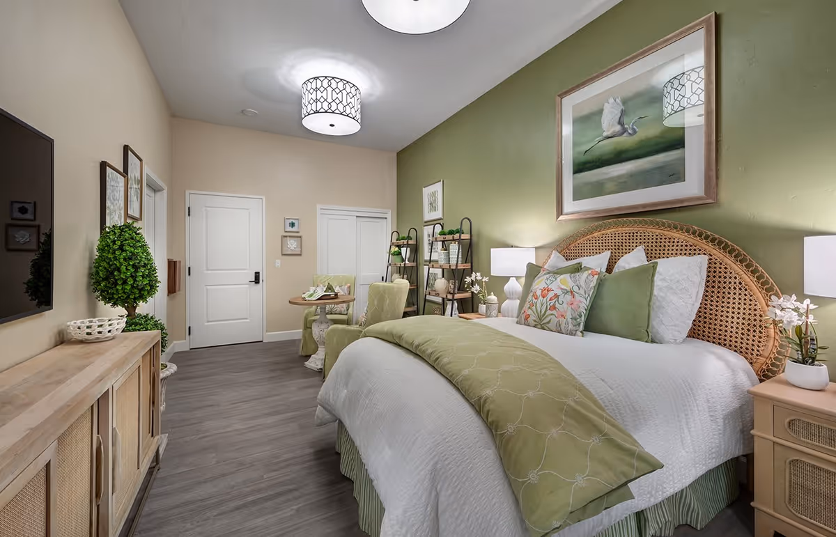 A cozy bedroom with a large bed featuring a wicker headboard, white and green bedding, and floral pillows. The room has a green accent wall with a framed painting of a bird in flight. There are two bedside tables with lamps and small plants, a wooden cabinet with a TV mounted above it, and a small seating area with two green chairs and a round table. The floor is wood, and the ceiling has a decorative light fixture.