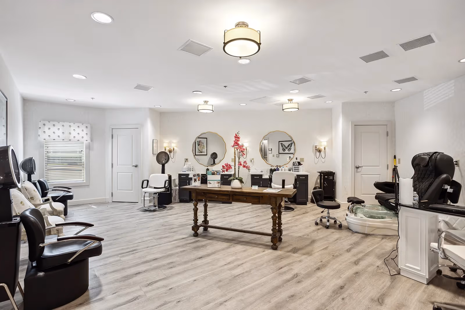 A spacious and well-lit beauty salon area in a senior living facility with multiple styling chairs, mirrors, and hair drying stations. The room features light-colored wood flooring, white walls, and ceiling lights. There is a wooden table with a vase of pink flowers in the center, and a pedicure chair with a foot bath on the right side.