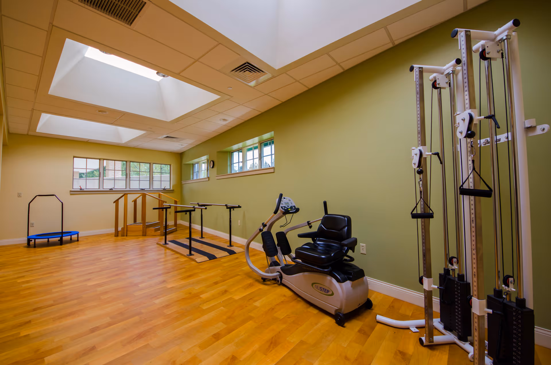 A well-lit exercise room with wooden flooring and light green walls. The room features various exercise equipment including a recumbent exercise bike, parallel bars for walking support, a small trampoline, and a multi-station weight machine. There are large windows and skylights allowing natural light to enter the space.