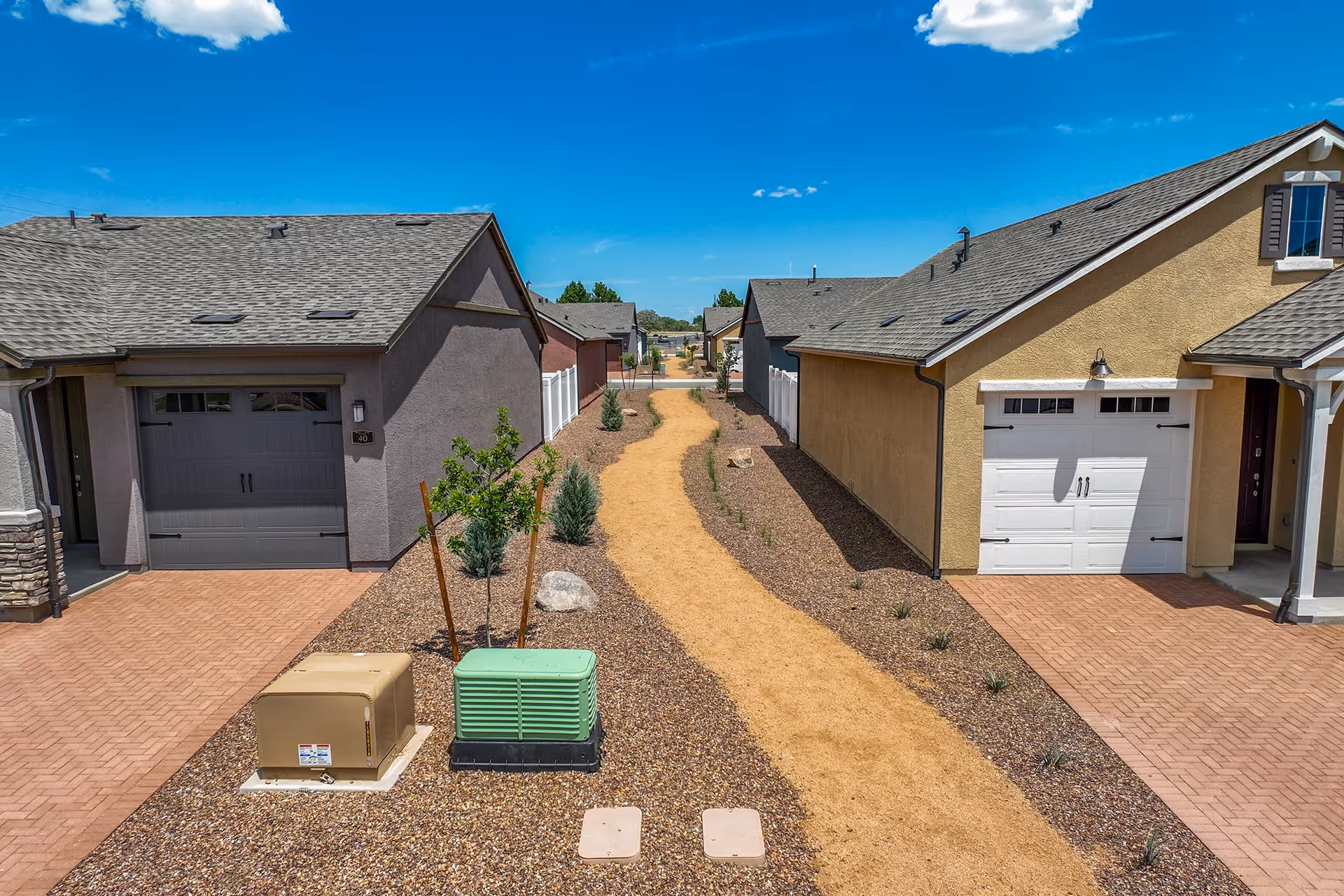 A narrow dirt walking path runs between two rows of single-story houses with garages, small trees, and shrubs planted along the sides under a clear blue sky.