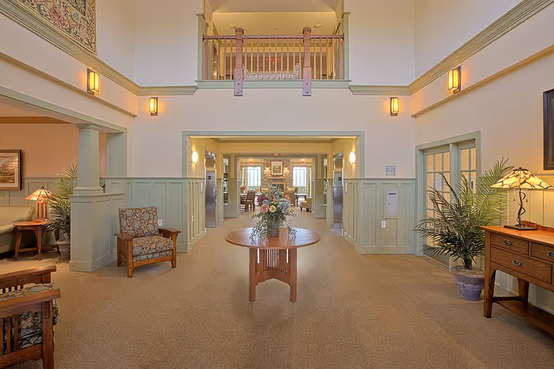 A spacious, well-lit interior common area with beige carpet and light green wainscoting. A round wooden table with a floral arrangement is centered in the hallway. There are wooden chairs with floral upholstery on the left and right sides, a wooden side table with a lamp and a potted plant on the right, and a balcony railing above the hallway entrance. The hallway leads to a room with more seating and a stone fireplace.