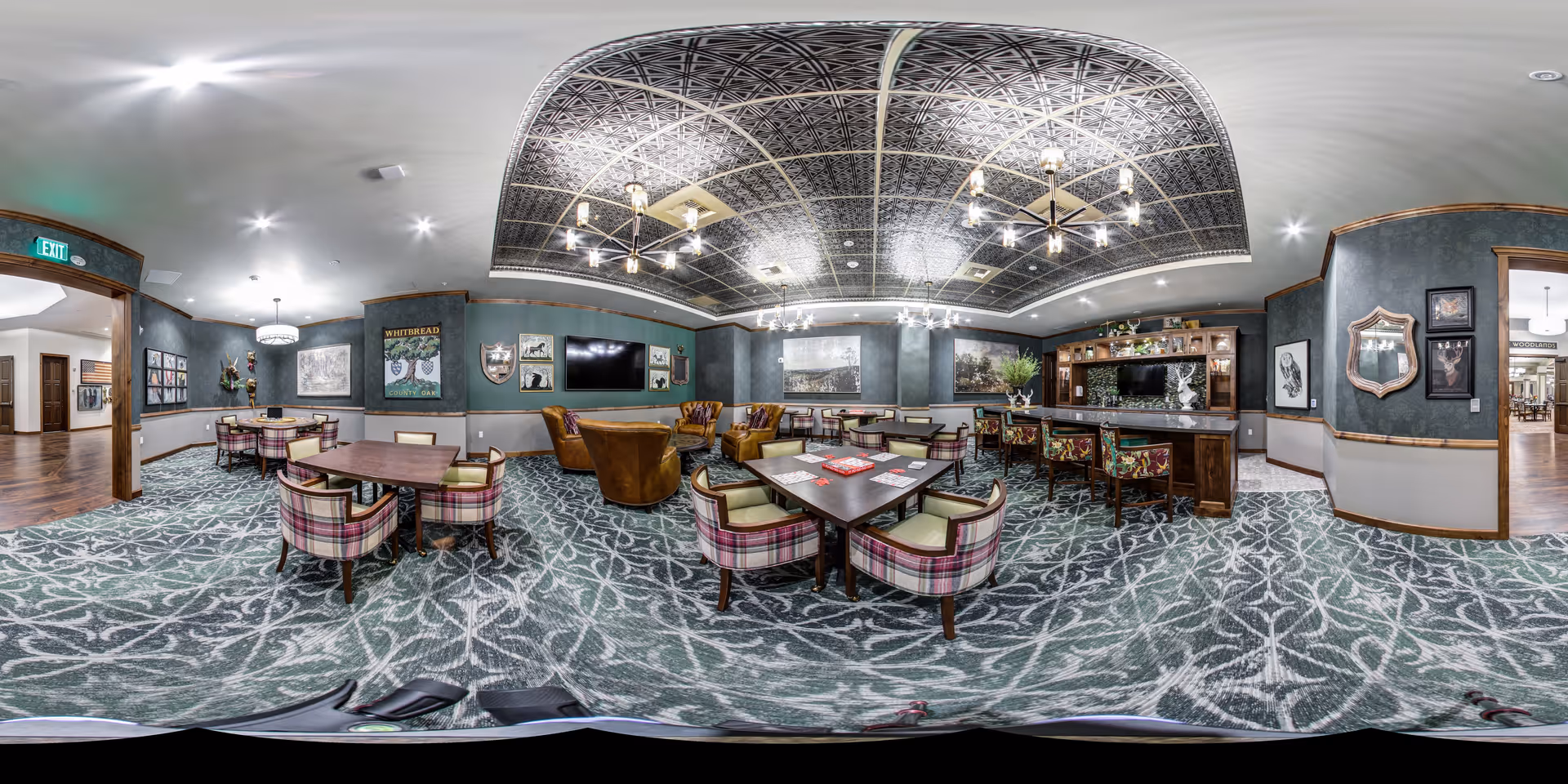A spacious, elegantly decorated common area with patterned carpet and a decorative ceiling. The room features several tables with plaid upholstered chairs, a seating area with brown leather armchairs, and a bar with high stools. The walls are adorned with framed pictures and artwork, and there is a large flat-screen TV mounted on one wall. The lighting includes modern chandeliers and recessed ceiling lights. The space appears warm and inviting, suitable for social gatherings or relaxation.
