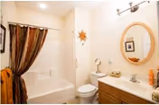 A clean bathroom featuring a white bathtub with a brown and orange striped shower curtain, a white toilet, a wooden vanity with a white countertop, a round mirror above the sink, a wall-mounted light fixture, and a small decorative sun hanging on the wall.