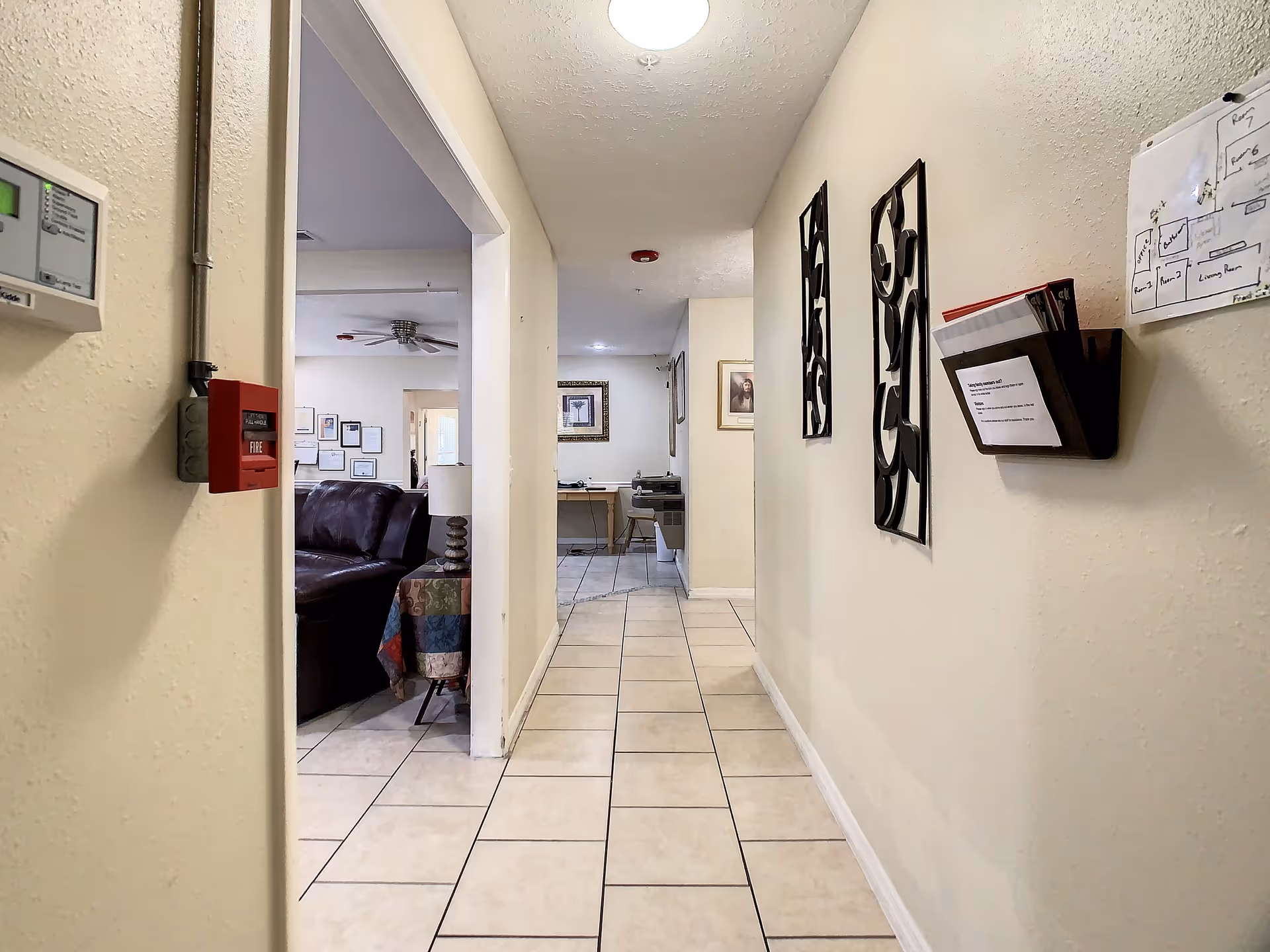 A tiled hallway in a senior living facility with beige walls and ceiling lights. On the left side, there is an open doorway leading to a room with a brown leather couch, a table with a colorful tablecloth, and a lamp. The hallway walls have decorative metal wall art and a wall-mounted organizer with papers. A fire alarm pull station and a thermostat are visible on the left wall near the doorway.