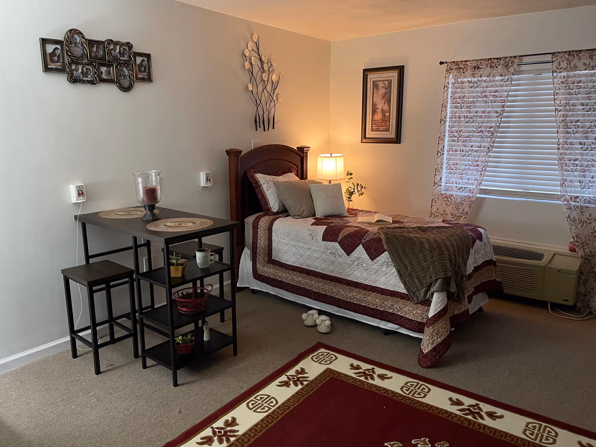 A cozy bedroom with a single bed covered in a quilted bedspread in shades of white, burgundy, and brown. The bed has multiple pillows and a folded blanket at the foot. Next to the bed is a small nightstand with a lamp and a framed picture on the wall above it. A window with floral curtains and blinds is on the right side. On the left side, there is a small black table with two stools and shelves holding decorative items and a candle. The floor has a red and beige patterned rug, and slippers are placed near the bed.
