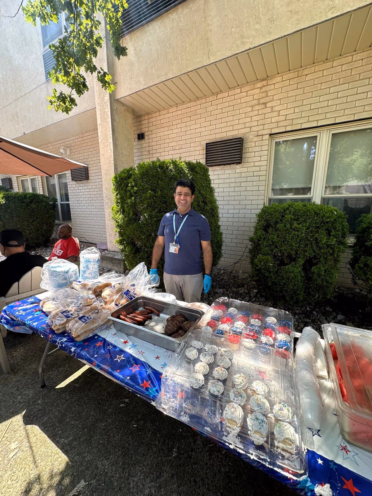 A man wearing a blue shirt and gloves stands behind a table covered with a patriotic tablecloth outdoors. The table has trays of hot dogs, hamburger patties, buns, cupcakes with white frosting, and other food items. There are bushes and a building with windows in the background, and two other people are seated nearby.