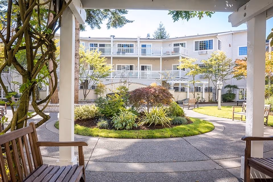 View of a landscaped outdoor courtyard at Weatherly Inn Tacoma, featuring a circular garden with various plants and small trees, surrounded by a paved walkway. Wooden benches are placed under a pergola with climbing vines, and a multi-story building with balconies and white railings is visible in the background.