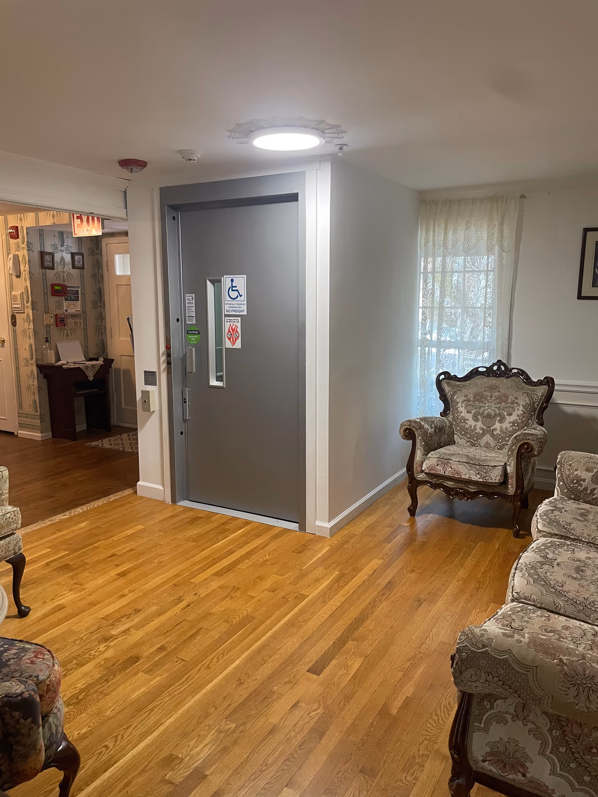 Interior view of a senior living facility with wooden flooring, vintage upholstered armchairs, a gray elevator door with accessibility signs, and a window with lace curtains allowing natural light.