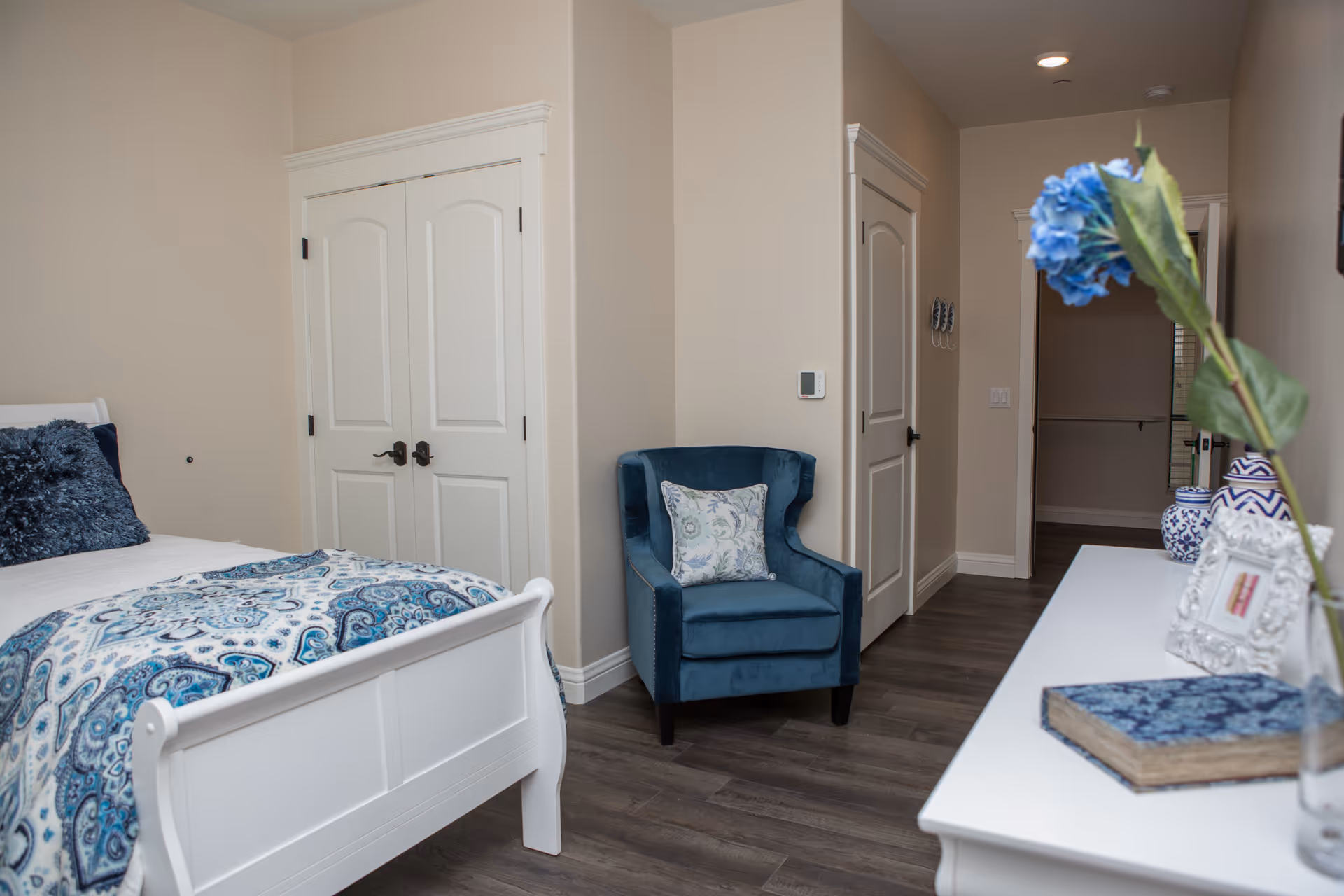 A cozy bedroom with a white wooden bed featuring blue and white patterned bedding and a dark blue pillow. There is a blue upholstered armchair with a floral cushion next to a beige wall. A white dresser with decorative items including a blue flower in a vase, a framed picture, and a book is visible. The room has wooden flooring and white doors, with a hallway leading to another room.
