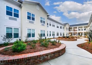 Courtyard with curved concrete walkway, brick planters, and a two-story white senior living building under a blue sky.