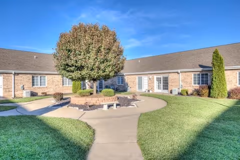Outdoor courtyard area of a senior living facility with a central tree surrounded by a circular brick planter and benches. The courtyard is bordered by a single-story brick building with multiple windows and doors. The sky is clear and blue.