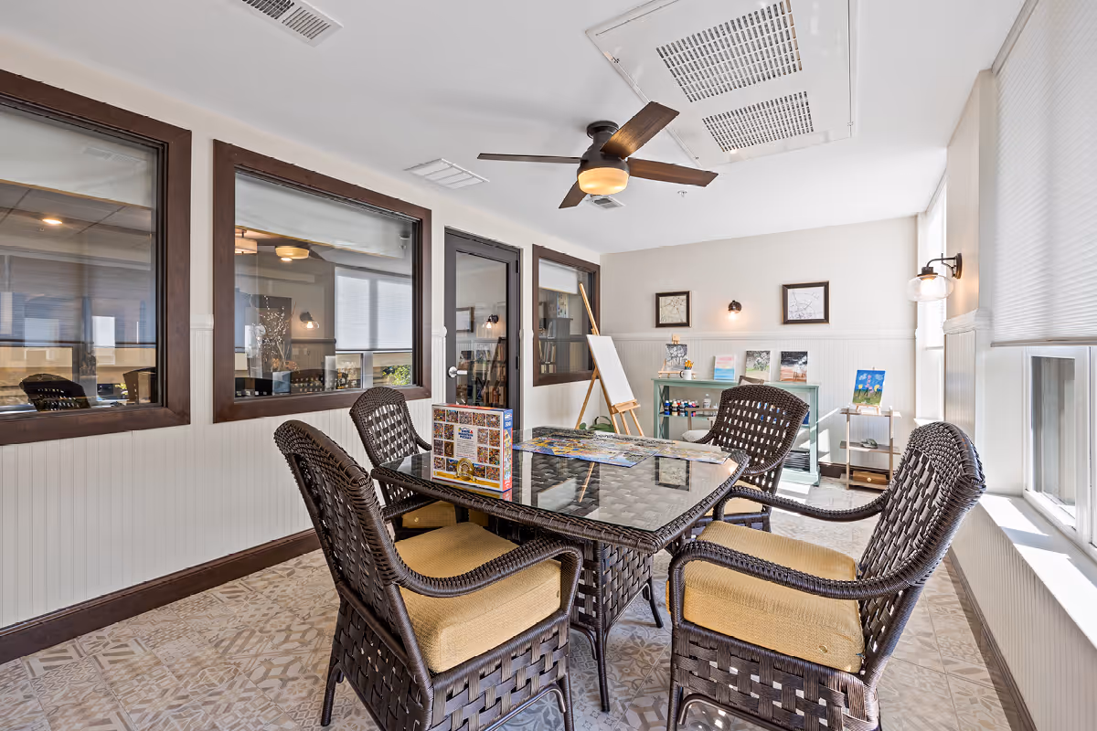 Bright activity room with a glass-top table surrounded by four wicker chairs, interior windows, an easel, and shelving with art supplies.