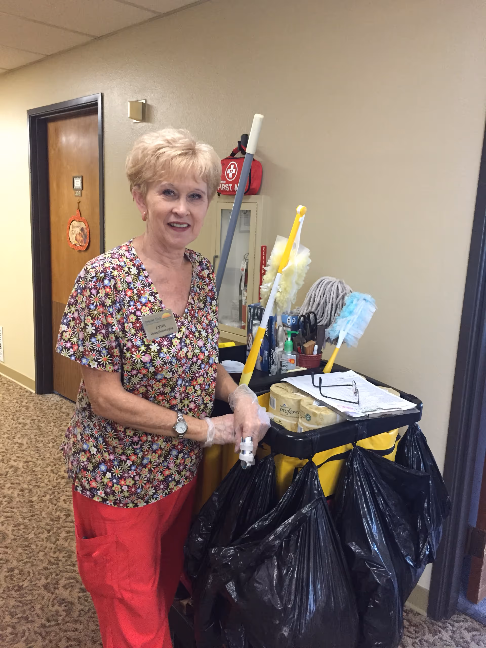 A woman wearing a floral patterned shirt and red pants stands in a hallway next to a cleaning cart filled with cleaning supplies and black trash bags. She is wearing gloves and smiling at the camera. Behind her is a door with a small decoration and a first aid kit mounted on the wall.